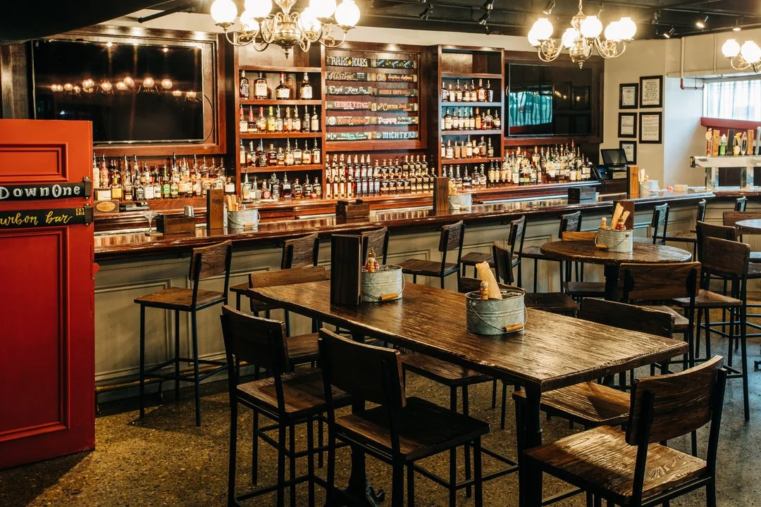 Warm, inviting bar with wooden tables, high chairs, and shelves stocked with bottles. A red door sign reads 'Down One Bourbon Bar,' suggesting a bourbon-focused establishment. The setting is cozy and ready for guests.