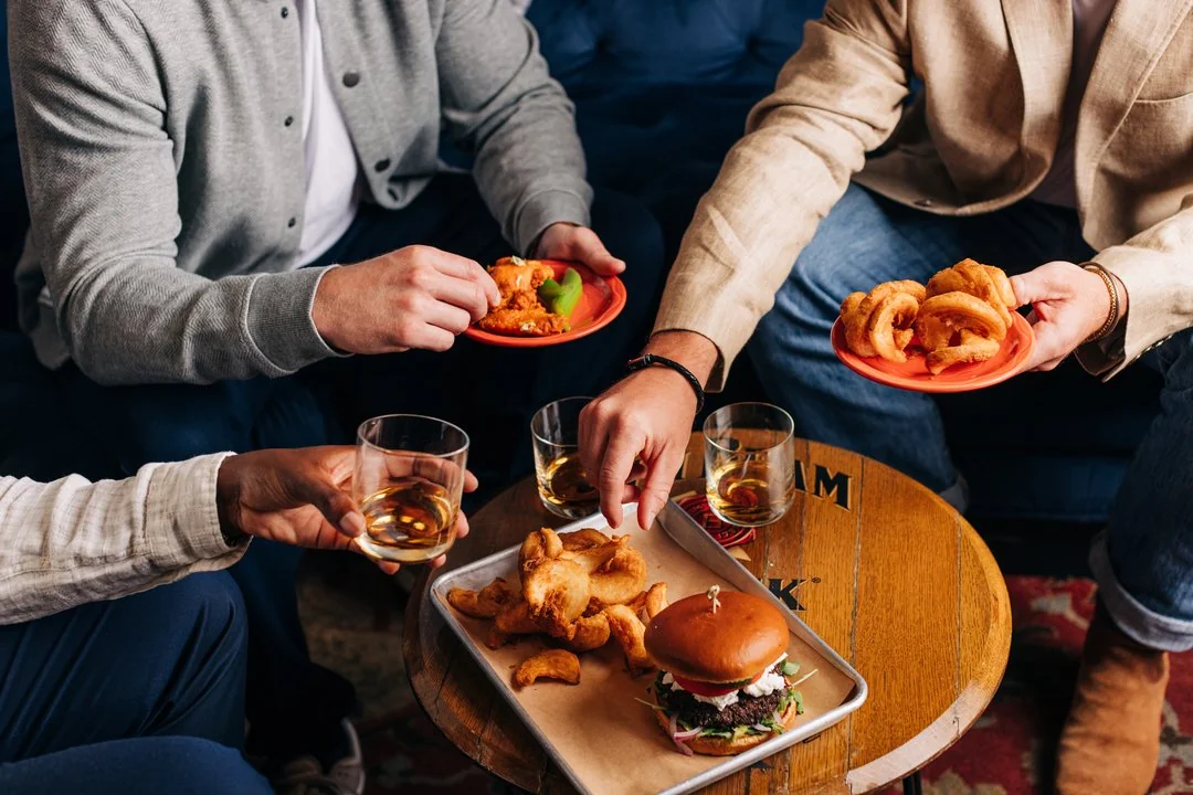 Three people sitting together share plates of fried snacks and drinks on a table, including a burger, fries, onion rings, and whiskey, suggesting a casual social gathering.