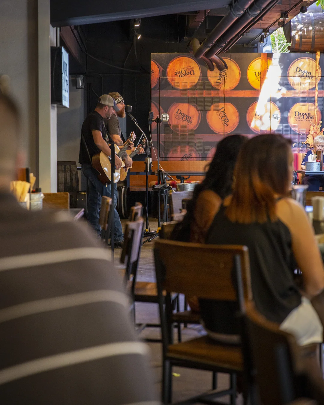 Two musicians perform with guitars at an indoor patio or bar area. Several people are seated and watching, with wooden barrels displayed on the wall in the background.
